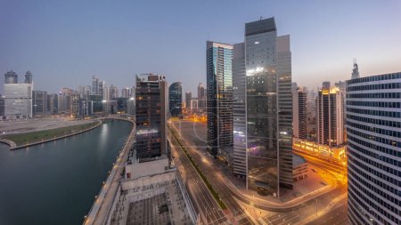 Photo for Cityscape of skyscrapers in Dubai Business Bay with water canal aerial night to day transition timelapse. Modern skyline with illuminated towers and road traffic before sunrise - Royalty Free Image