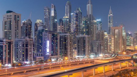 Téléchargez les photos : Dubai Marina plus haut bloc de gratte-ciel jour à nuit passage timelapse. Vue aérienne du quartier JLT vers les immeubles à appartements, les hôtels et les tours de bureaux près de l'autoroute. - en image libre de droit