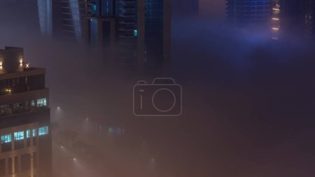 Foto de Buildings are covered in thick layer of fog in Business Bay night . Illuminated skyscrapers around water canal with traffic on a road aerial top view - Imagen libre de derechos