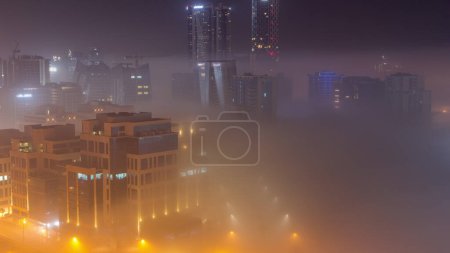 Téléchargez les photos : Buildings are covered in thick layer of fog in Business Bay night . Illuminated skyscrapers around water canal aerial top view - en image libre de droit