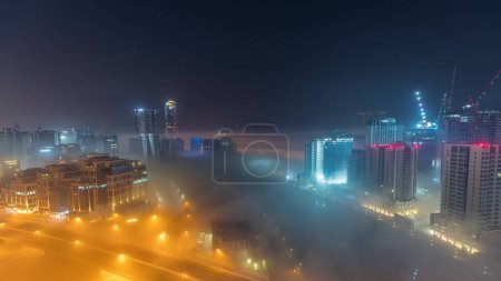 Téléchargez les photos : Buildings are covered in thick layer of fog in Business Bay during all night . Illuminated skyscrapers around water canal aerial top view - en image libre de droit