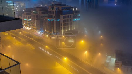 Foto de Buildings are covered in thick layer of fog in Business Bay night . Illuminated office buildings with traffic on a road aerial top view - Imagen libre de derechos