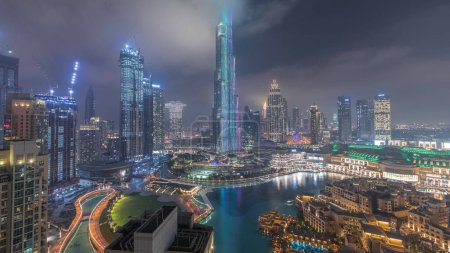 Photo for Skyscrapers rising above Dubai downtown night , mall and fountain surrounded by illuminated modern buildings aerial top panoramic view with cloudy sky - Royalty Free Image