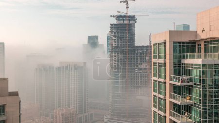 Téléchargez les photos : Dubai skyscrapers covered by morning fog in business bay district with construction site during sunrise . Aerial view from top of downtown in United Arab Emirates. - en image libre de droit