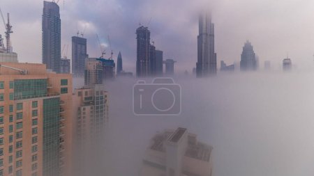 Téléchargez les photos : Aerial view of Dubai city early morning during fog . Sunrise at futuristic city skyline with skyscrapers and towers from above. Sun reflected from glass surface with rays of light - en image libre de droit