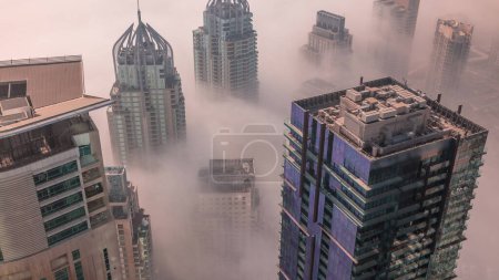 Foto de Towers covered by rare early morning winter fog above the Dubai Marina skyline and skyscrapers rooftops aerial . Top view from above clouds. Dubai, UAE - Imagen libre de derechos