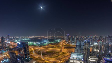 Téléchargez les photos : Huge highway crossroad junction panorama between JLT district and Dubai Marina intersected by Sheikh Zayed Road aerial  during all night with rising moon. Golf course near illuminated towers and skyscrapers with busy traffic - en image libre de droit