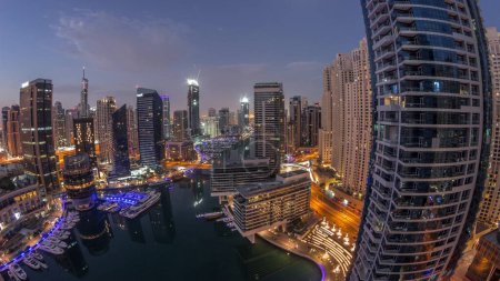Téléchargez les photos : Aerial view to Dubai marina illuminated skyscrapers around canal with floating yachts night to day transition  panorama. White boats are parked in yacht club before sunrise - en image libre de droit