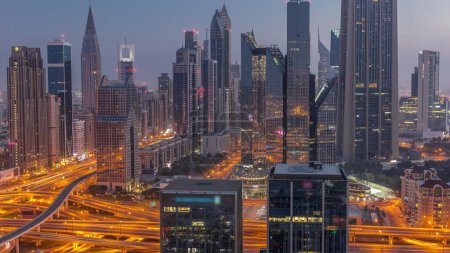Foto de Panorama of Dubai Financial Center district with tall skyscrapers with illumination night to day transition . Aerial view to towers along busy highway before sunrise - Imagen libre de derechos