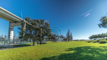 Foto de The Vasco da Gama Bridge timelapse hyperlapse viewed from green lawn with trees. Cable-stayed longest bridge flanked by viaducts and rangeviews that spans Tagus River in Park Nations. Lisbon, Portugal - Imagen libre de derechos