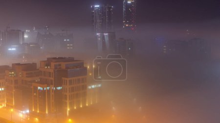 Foto de Los edificios están cubiertos de una gruesa capa de niebla en el lapso de tiempo nocturno de Business Bay. Rascacielos iluminados alrededor del canal de agua vista aérea - Imagen libre de derechos