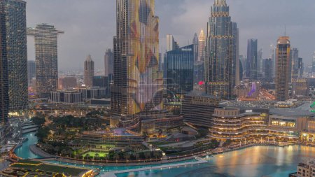 Téléchargez les photos : Gratte-ciel s'élevant au-dessus du centre-ville de Dubaï de jour à la nuit transition timelapse, centre commercial et fontaine entourés de bâtiments modernes vue aérienne avec ciel nuageux coloré - en image libre de droit