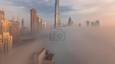 Foto de Vista panorámica aérea de la ciudad de Dubai temprano en la mañana durante el timelapse niebla. Salida del sol en el horizonte futurista de la ciudad con rascacielos y torres desde arriba. Sol reflejado desde la superficie de vidrio - Imagen libre de derechos