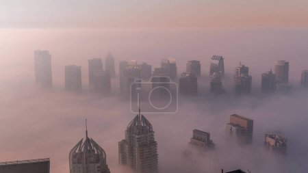 Téléchargez les photos : Le brouillard couvrait les gratte-ciels et les tours de marina de la JLT près de Sheikh Zayed Road pendant le lever du soleil. Immeuble résidentiel matin brumeux avec lumière chaude colorée - en image libre de droit