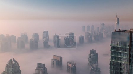 Téléchargez les photos : Le brouillard couvrait les gratte-ciel et les tours de marina de la JLT près de Sheikh Zayed Road après le lever du soleil. Immeuble résidentiel matin brumeux avec lumière orange chaude - en image libre de droit