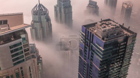 Photo for Towers covered by rare early morning winter fog above the Dubai Marina skyline and skyscrapers rooftops aerial timelapse. Top view from above clouds. Dubai, UAE - Royalty Free Image