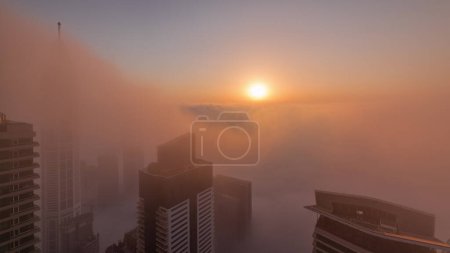 Foto de Salida del sol sobre la rara niebla invernal de la madrugada sobre el horizonte de Dubai Marina y los techos de rascacielos timelapse aéreo. Vista superior desde las nubes superiores. Dubai, Emiratos Árabes Unidos - Imagen libre de derechos