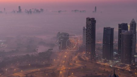 Foto de Gran cruce de caminos entre el distrito JLT y el puerto deportivo de Dubái intersecado por Sheikh Zayed Road noche a día el lapso de tiempo aéreo de transición. Tráfico de coches cubierto por niebla matutina - Imagen libre de derechos