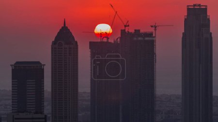 Téléchargez les photos : Skyline avec l'architecture moderne des tours de la baie d'affaires de Dubaï au coucher du soleil timelapse avec grand soleil rouge. Vue aérienne rapprochée avec canal et chantier - en image libre de droit