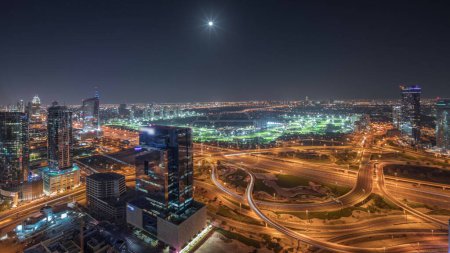 Téléchargez les photos : Panorama montrant la ville des médias, la marina de Dubaï et le JLT illuminaient les gratte-ciel le long de Sheikh Zayed Road avec une grande nuit aérienne à la jonction du carrefour. Lune montante sur les immeubles résidentiels et de bureaux et le golf - en image libre de droit