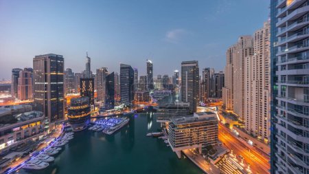 Téléchargez les photos : Vue panoramique aérienne sur la marina de Dubaï illuminait les gratte-ciel autour du canal avec des yachts flottants nuit à jour passage timelapse. Les bateaux blancs sont garés dans le yacht club avant le lever du soleil - en image libre de droit