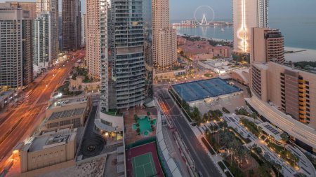Téléchargez les photos : Vue panoramique sur le port de plaisance de Dubaï et la région de JBR et la célèbre roue Ferris nuit aérienne à la transition de jour timelapse. gratte-ciel lumineux au lever du soleil avec de longues ombres - en image libre de droit