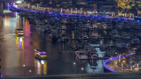 Téléchargez les photos : De nombreux yachts et bateaux sont stationnés dans le timelapse de nuit aérienne du port. Dubai Marina front de mer zone de marche. Bateau moderne monte le ruisseau sous le pont avec la circulation - en image libre de droit
