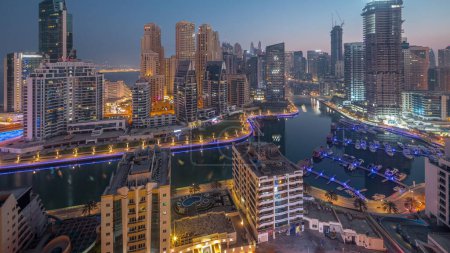 Foto de Dubai Marina con barcos y yates estacionados en el puerto y rascacielos iluminados alrededor del canal aéreo noche a día timelapse transición antes del amanecer. Torres del distrito JBR en segundo plano - Imagen libre de derechos