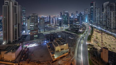 Téléchargez les photos : Panorama montrant Bay Avenue avec des tours modernes éclairées développement résidentiel dans Business Bay ciel nocturne timelapse, Dubaï, Émirats arabes unis. gratte-ciel avec circulation sur une route près d'un grand parking - en image libre de droit