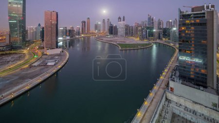 Téléchargez les photos : Paysage urbain de gratte-ciel dans la baie d'affaires de Dubaï avec canal d'eau ciel nuit à jour transition timelapse avec la lune couchant. Skyline moderne avec tours éclairées et front de mer avant le lever du soleil - en image libre de droit