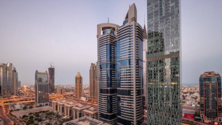 Téléchargez les photos : Vue aérienne du quartier financier international de Dubaï avec de nombreux gratte-ciels nuit à jour passage timelapse. Trafic sur une route près d'un parking avec des tours éclairées. Dubai, Émirats arabes unis. - en image libre de droit