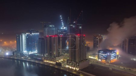 Foto de Los edificios están cubiertos de una gruesa capa de niebla en la noche de Business Bay. Rascacielos iluminados cerca del canal de agua con vista aérea del sitio de construcción - Imagen libre de derechos