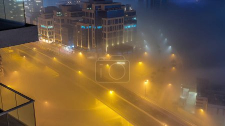 Téléchargez les photos : Buildings are covered in thick layer of fog in Business Bay night . Illuminated office buildings with traffic on a road aerial top view - en image libre de droit