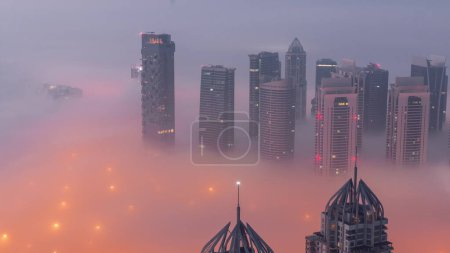 Foto de JLT skyscrapers near Sheikh Zayed Road aerial night to day transition . Illuminated residential buildings and traffic on crossroad junction. Foggy morning before sunrise - Imagen libre de derechos