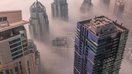 Foto de Towers covered by rare early morning winter fog above the Dubai Marina skyline and skyscrapers rooftops aerial . Top view from above clouds. Dubai, UAE - Imagen libre de derechos