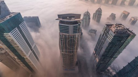 Téléchargez les photos : Towers covered by rare early morning winter fog between the Dubai Marina skyline and skyscrapers rooftops aerial . Look down view from above clouds. Dubai, UAE - en image libre de droit