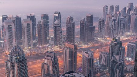 Photo for JLT skyscrapers and marina towers near Sheikh Zayed Road aerial night to day transition . Illuminated residential buildings and skyline with villas. Foggy morning before sunrise - Royalty Free Image