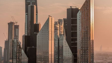 Foto de Sol reflejado desde torres de oficinas superficie de vidrio en el distrito financiero de la ciudad de Dubai. Vista aérea con el sitio de construcción detrás durante la puesta del sol con luz naranja - Imagen libre de derechos