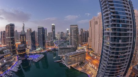 Foto de Aerial view to Dubai marina illuminated skyscrapers around canal with floating yachts night to day transition  panorama. White boats are parked in yacht club before sunrise - Imagen libre de derechos