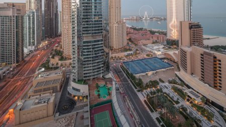 Téléchargez les photos : Panoramic view of the Dubai Marina and JBR area and the famous Ferris Wheel aerial night to day transition . Illuminated skyscrapers during sunrise with long shadows - en image libre de droit