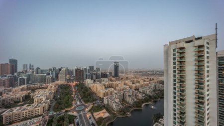 Téléchargez les photos : Des gratte-ciels dans le quartier de Barsha Heights et des immeubles de faible hauteur dans le quartier des Verts transition aérienne de jour à nuit. Skyline de Dubaï avec palmiers et fontaine sur étang - en image libre de droit