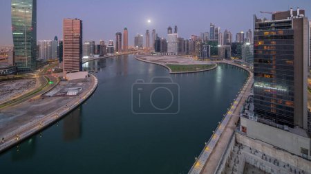 Téléchargez les photos : Cityscape of skyscrapers in Dubai Business Bay with water canal aerial night to day transition  with Moon setting down. Modern skyline with illuminated towers and waterfront before sunrise - en image libre de droit