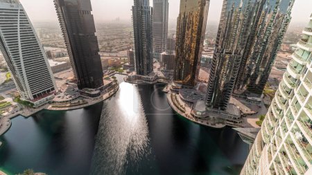 Téléchargez les photos : Grands bâtiments résidentiels à l'antenne du quartier JLT, qui fait partie du quartier mixte du centre multi-commodités de Dubaï au lever du soleil. Soleil réfléchi par une eau dans un étang - en image libre de droit