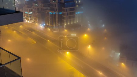 Téléchargez les photos : Buildings are covered in thick layer of fog in Business Bay night . Illuminated office buildings with traffic on a road aerial top view - en image libre de droit