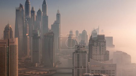 Foto de Vista de varios rascacielos en el bloque recidencial más alto de Dubai Marina durante la salida del sol aérea con canal artificial. Muchas torres y yates cubiertos por la niebla de la mañana - Imagen libre de derechos