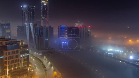Foto de Buildings are covered in thick layer of fog in Business Bay night . Illuminated skyscrapers around water canal aerial panoramic view - Imagen libre de derechos