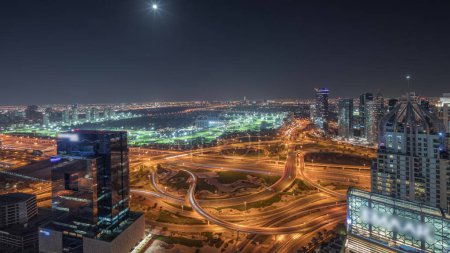 Foto de Panorama que muestra la ciudad de los medios de comunicación, el puerto deportivo de Dubái y los rascacielos iluminados JLT a lo largo de Sheikh Zayed Road con una gran noche aérea de cruce. Luna naciente sobre edificios residenciales y de oficinas y golf - Imagen libre de derechos