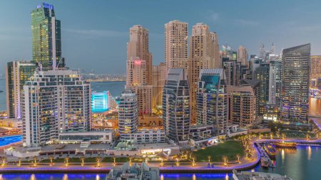 Foto de Dubai Marina con varios barcos cerca de césped verde en el paseo marítimo y rascacielos alrededor de la antena del canal día a la transición nocturna. Torres del distrito JBR en segundo plano - Imagen libre de derechos