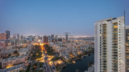 Foto de Rascacielos en el distrito de Barsha Heights y edificios de poca altura en el distrito de Greens transición aérea día a noche. Dubai skyline con palmeras y fuente en el estanque - Imagen libre de derechos