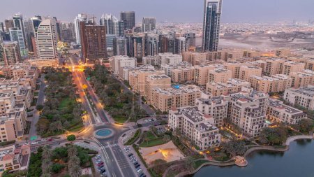 Téléchargez les photos : Bâtiments de faible hauteur dans le quartier des Verts et gratte-ciel dans le quartier de Barsha Heights transition aérienne du jour à la nuit panoramique. Skyline de Dubaï avec palmiers et arbres - en image libre de droit